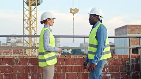 Multiethnic-man-and-woman-in-hardhats-standing-at-the-construcing-site,-talking-and-shaking-hands.-Builder-and-architect-meeting.-Outdoors.